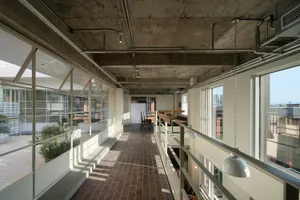 Long perspective view of an industrial-style mezzanine corridor featuring exposed concrete ceiling, large steel-framed windows, brick flooring, and white pendant lights overlooking an open loft space.