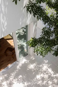 High-angle view of a modern courtyard featuring white walls, light gray tiled flooring, wooden flooring inside, a wooden chair, and lush green foliage casting dramatic leaf shadows.