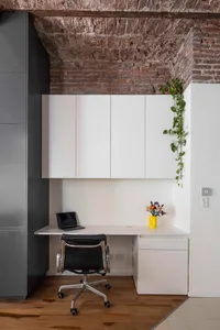 Modern home office nook featuring a white built-in desk, black leather office chair, exposed brick ceiling, and trailing green plants.