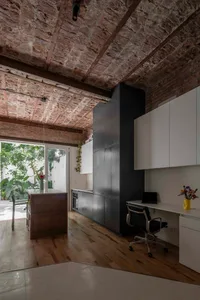 Industrial loft interior featuring exposed brick ceiling, dark grey kitchen cabinetry, white office desk with black leather chair, and large glass doors opening to a sunny garden.