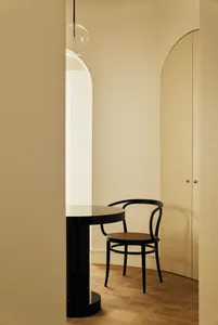 Modern minimalist entryway corner with a black bentwood chair, glossy black round pedestal table, arched doorway, and warm beige walls under a glass globe pendant light.