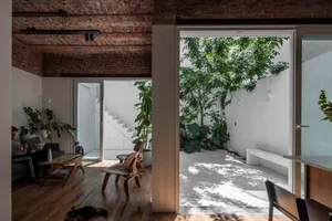 Modern loft interior with exposed brick ceiling, wooden floors, Eames-style chairs, and a large open courtyard filled with trees and plants, with a dog resting on the floor and a person sitting on a grey sofa.