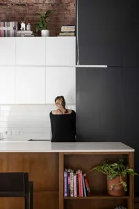 Modern industrial-style kitchen featuring exposed red brick wall, white flat-panel cabinets, white tiled backsplash, walnut wood open shelving, and a white countertop island.