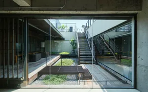 View from inside a modern concrete house looking out onto a sunlit courtyard with wooden deck, gravel garden, potted plants, and external staircase.