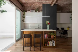 Modern industrial loft kitchen with wooden central island, two dark bar stools, white cabinetry, exposed brick ceiling, and large glass doors opening to a courtyard.
