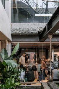 People gathering at a modern industrial-style cafe featuring exposed concrete, large glass walls, steel beams, and abundant indoor plants under a glass atrium.