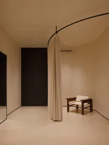 Minimalist interior corner featuring a walnut wood armchair with off-white upholstery, positioned beside a curved black rail holding a heavy cream fabric curtain that acts as a soft room divider.
