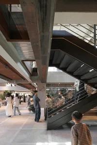 People standing in a spacious multi-level brutalist atrium featuring massive exposed concrete beams and a dramatic dark metal staircase.