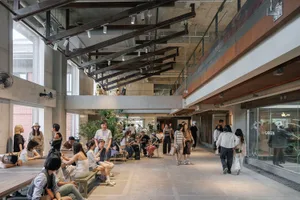 Busy multi-level industrial-style public space with exposed concrete, dark steel beams, large windows, and people sitting at long wooden tables and walking through the atrium.