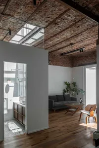 Open-plan loft living room featuring exposed brick ceiling, large skylight, grey sofa, wooden Eames-style chair, and adjacent white-tiled bathroom with terrazzo floor.