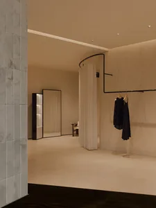 Modern minimalist dressing room featuring raw micro-cement walls, a curved black clothing rail with hanging garments, a tall mirror, and soft recessed lighting.