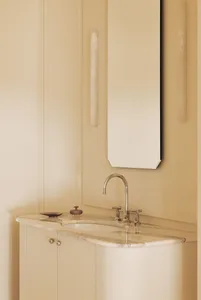 Close-up of a bathroom vanity with white and beige marble countertop, integrated sink, polished chrome faucet, and a tall rectangular mirror against cream-colored paneled walls.