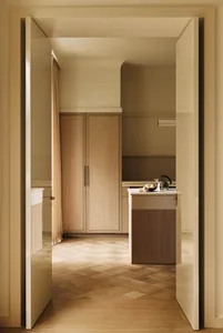 View through open white double doors into a minimalist butler pantry featuring light oak cabinetry, a marble-topped island with a kettle and fruit bowl, and warm herringbone parquet flooring.