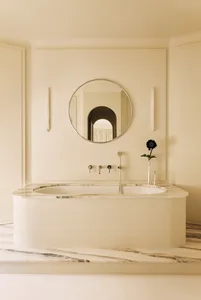 Freestanding curved marble bathtub with chrome freestanding faucet, round mirror, and single black sunflower in a minimalist all-white marble bathroom