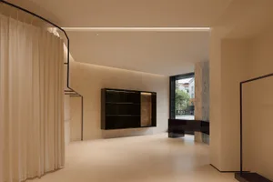 Modern minimalist living room featuring travertine stone walls, a floating dark wood shelving unit with integrated lighting, sheer curtains on a curved black rail, and a view to an outdoor courtyard.