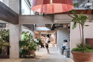 Modern industrial-style cafe interior featuring exposed concrete beams, large red pendant lamp, abundant potted plants, and people relaxing in a bright open space.