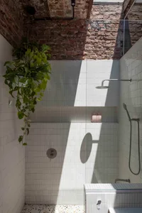 Modern shower space with white tiled walls, exposed red brick upper wall, hanging potted plants, chrome rainfall shower head, and terrazzo floor under bright sunlight.