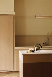 Modern minimalist kitchen featuring light oak cabinetry, a travertine countertop and backsplash, a stainless steel kettle, and fresh onions on the island.