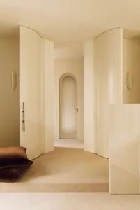Minimalist curved hallway in warm cream tones featuring built-in arched doorway, wall sconces, and two large brown velvet floor cushions in the foreground.