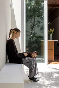 A woman with braided hair sits on a white built-in window seat, gently holding a Siamese cat on her lap while sunlight streams through a large glass door overlooking green trees.