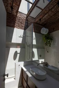 Modern industrial loft bathroom featuring white subway tile walls, two vessel sinks on a concrete countertop, exposed brick ceiling with large skylights, and strong natural light creating dramatic shadows.