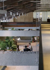 View from upper level looking down into a double-height industrial cafe with exposed concrete beams, wooden ceiling, abundant indoor plants, and people sitting at tables on the ground floor.