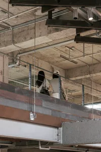 View looking up at an industrial-style mezzanine with exposed concrete ceiling, steel beams, cable railings, and two women walking on the upper level.