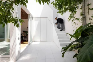 Bright white modern courtyard interior with floating concrete staircase, large green plants, glass sliding doors, and two people with a cat on the stairs.