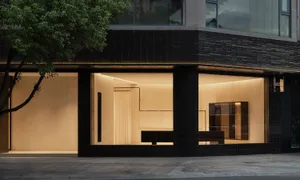 Modern minimalist luxury storefront at night with black textured tile facade, large glass windows revealing warm beige interiors and custom wooden display fixtures.