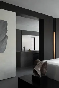 Modern minimalist living room featuring dark concrete walls, a large abstract stone sculpture on the left, sculptural bronze artwork on a black pedestal, and a view into a concrete kitchen with a sleek faucet.