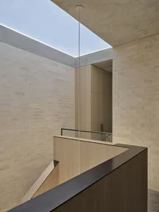 Modern minimalist atrium interior featuring light-colored brick walls, light wood staircase railing, concrete ceiling, and a large skylight allowing natural daylight to flood the space.