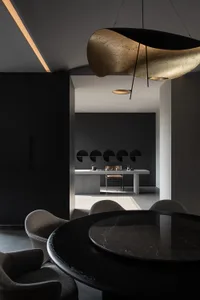Modern dark minimalist dining area featuring a round black marble table, sculptural gold and black pendant light, textured boucle chairs, and a view into a dark kitchen with arched niches.