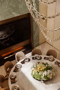 High-angle view of a luxurious round white marble dining table set for six with neutral upholstered chairs, a large floral arrangement, modern fireplace with green marble surround, and a dramatic bubble chandelier.