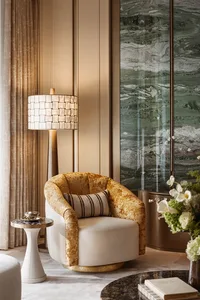 Luxurious living room corner with a golden velvet and cream swivel armchair, striped lumbar pillow, tall floor lamp with mother-of-pearl shade, green marble wall panels, and a white side table.