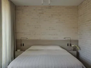Modern minimalist bedroom with light beige exposed brick wall, wooden headboard, natural linen bedding, wall-mounted reading lamps, and sheer curtains.