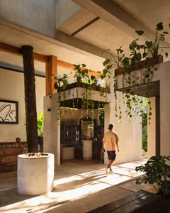 A man walking through a sunlit tropical modern indoor-outdoor patio featuring concrete architecture, warm wood beams, abundant hanging plants, and a cylindrical concrete fire pit.
