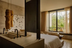 Modern bedroom interior featuring a raw concrete double vanity with black faucets, woven rattan pendant lights, dark wood partition, and large windows overlooking tropical greenery.