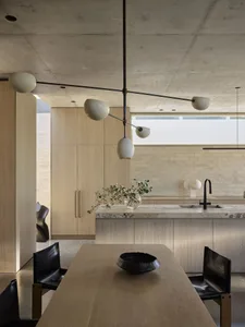 Modern minimalist kitchen and dining area featuring light oak cabinetry, a large marble kitchen island, black leather dining chairs, and a dramatic black metal and white glass pendant light against a raw concrete ceiling.