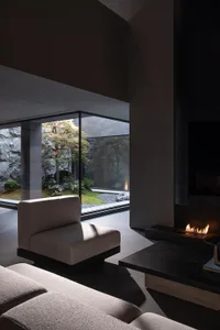 Modern minimalist living room interior featuring raw concrete walls, a built-in linear fireplace, low-profile beige fabric sofa, and a tranquil Japanese garden visible through floor-to-ceiling glass windows.