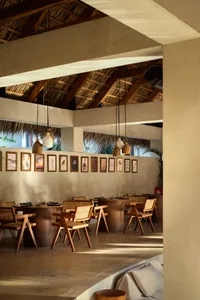 開放式熱帶餐廳室內空間，設有柚木圓桌、藤編餐椅、茅草屋頂，以及一面掛滿植物與風景畫作的肌理混凝土牆。