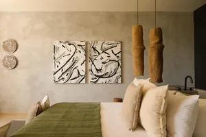 Modern wabi-sabi bedroom featuring a concrete wall, two large abstract black and white paintings, olive green herringbone throw blanket, cream linen pillows, and two tall cylindrical woven rattan pendant lights.