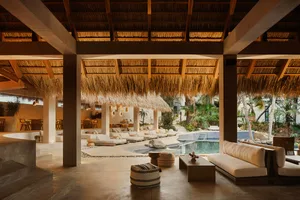 Spacious open-air lounge in a tropical villa featuring concrete pillars, thatched roof, cream-colored sofas, woven poufs, and an infinity pool overlooking lush palm trees.