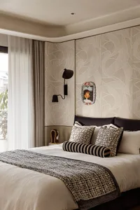 Modern bedroom interior with beige leaf-patterned textured wall panels, dark leather headboard, black wall sconce, and bed dressed in white linen with black and beige geometric patterned pillows and throw.