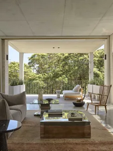 Modern minimalist living area opening to a covered patio with concrete ceiling, steel and glass coffee tables, wooden lounge chair, and dense forest view beyond the railing.