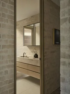 View into a modern brutalist bathroom with light travertine floating vanity, oak drawers, black matte faucet, and textured brick walls.