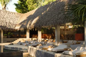 Tropical outdoor poolside lounge with multiple white cushioned daybeds, rolled towels, wooden bowls, and large thatched-roof structures in warm sunlight surrounded by lush greenery.