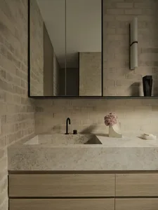 Modern bathroom featuring a solid travertine floating vanity with integrated sink, matte black faucet, light wood cabinetry, and textured beige brick walls with a large frameless mirror.