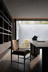 Modern minimalist home office featuring a dark walnut desk, brown leather chair, built-in shelving, and a large window overlooking a tranquil garden with stone wall.