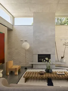 Modern minimalist living room featuring a large concrete fireplace wall, oak lattice coffee table on a cream rug, wooden bench, paper floor lamp, and natural daylight from high windows.