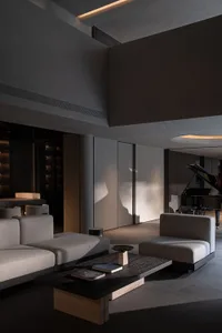 Modern minimalist double-height living room featuring raw concrete walls, low-profile grey sectional sofa, dark stone coffee table, and a black grand piano in the background.